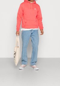 Korallfärgad hoodie med framficka och dragsnören, i kombination med ljusblå jeans. Canvas tote väska med grafiskt tryck. Vita sneakers med rosa detaljer.