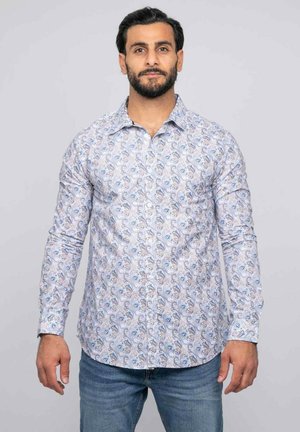 Homme aux cheveux foncés et à la barbe, portant une chemise blanche avec des motifs cachemire bleus et un jean bleu, debout devant un fond uni.