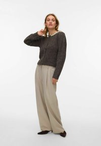 Tumši brūns adīts kardigans ar pērļu rotātu apkakli, kombinēts ar gaiši bēšām plašu kāju bikšu biksēm un tumši brūnām smailām kurpēm.