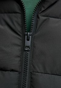 Svart polstret jakke med sentral glidelås og en merket glidelåstrekk, vist mot en teksturert grønn bakgrunn.