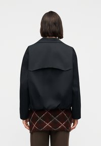 Άτομο με κοντά σκούρα μαλλιά, φορώντας μαύρο μπουφάν πάνω από κόκκινη και καφέ καρό φούστα, στέκεται πλάτη σε απλό φόντο.