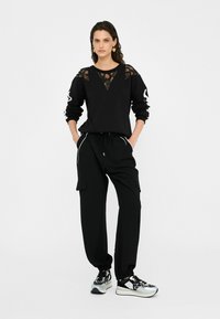 Sweatshirt noir avec empiècement en dentelle, pantalon cargo noir et baskets noir et argent. Coupe décontractée, taille élastique, poches latérales.