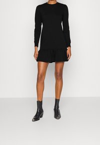 Black pullover dress with long sleeves, ruffled hem, and a round neckline, paired with black ankle boots featuring a silver toe detail.