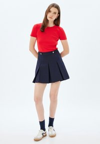 Camiseta roja ajustada de manga corta, combinada con una falda azul marino con pliegues, de cintura alta y dos botones plateados en la parte delantera.