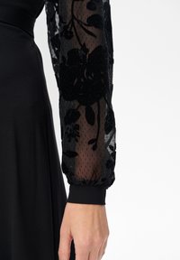 Schwarzes Kleid mit durchsichtigen, gemusterten Ärmeln mit floralem Applikation und einem figurbetonten Oberteil. Die Ärmelsäume haben ein gerafftes Bündchen.