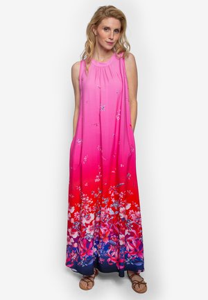 Vrouw staat in een mouwloze maxi-jurk met een roze naar rood ombre kleurverloop en bloemenprint, gecombineerd met sandalen, tegen een effen witte achtergrond.