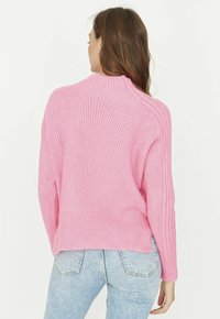 Maglione rosa lavorato a maglia con collo alto, trama a coste, spalle scese e spacchi laterali, abbinato a jeans azzurri.
