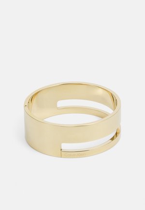 Armband van metaal met goudkleurige tint, brede band en twee horizontale uitsparingen, gegraveerd met het Calvin Klein-logo aan één kant.