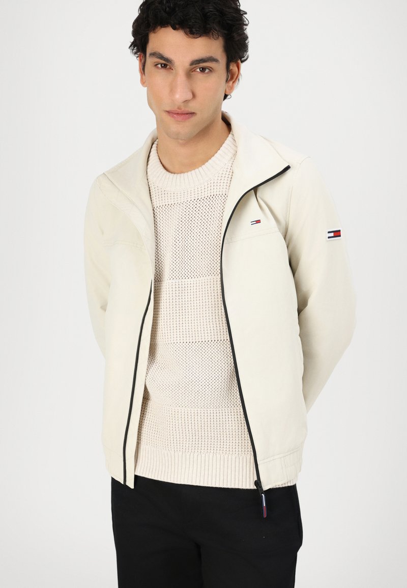 Chaqueta beige clara con cremallera y detalles en negro, que presenta un pequeño logo en el brazo izquierdo. Lleva puesta sobre un suéter texturizado color crema y pantalones negros.