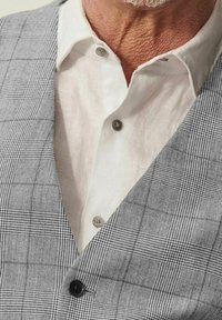 Gilet grigio a quadri con un motivo testurizzato e bottone nero, indossato sopra una camicia di lino bianca con bottoni grigio chiaro.