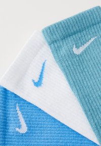 Trīs Nike zeķu pāri zili, gaiši zili un balti. Katram ir kontrastējoša Swoosh zīme ar ribotu tekstūru un dažādiem krāsu rakstiem.