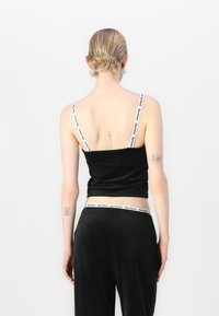 Crop top in velluto nero con spalline sottili che presenta un logo bianco di marca. La parte posteriore ha un collo a barca e una finitura liscia e testurizzata.