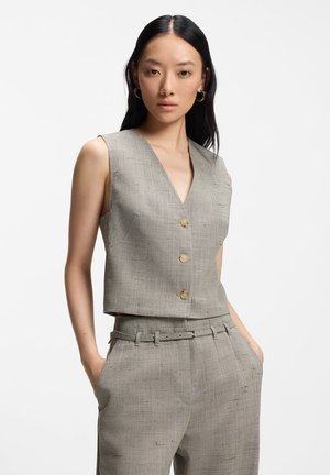 Nainen, jolla on yllään hihaton beige napitettava liivi ja siihen sopivat korkeavyötäröiset housut vyöllä, seisoo kädet taskuissa valkoista taustaa vasten.