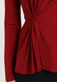 Rotes Langarmoberteil mit gerafften Falten an der Seite, aus weichem Jersey-Stoff mit glatter Textur und asymmetrischem Saum.