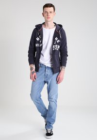 Navy-blaues Zip-Hoodie mit weißem Druck, getragen über einem schlichten weißen T-Shirt. Hellblaue Jeans und schwarze Sneakers mit weißen Streifen runden den Look ab.