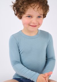 Camicia a maniche lunghe di un blu chiaro, realizzata in cotone, con scollatura rotonda e trama a costine, indossata da un bambino seduto con le mani unite.