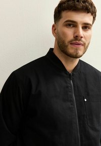 Jonge man met kort krullend haar en baard, gekleed in een zwarte zip-up jas met borstzak, staand tegen een effen lichte muur.