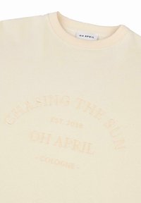 Sudadera color crema con bordado en relieve que presenta el texto "CHASING THE SUN EST. 2018 OH APRIL COLOGNE" en un diseño circular.