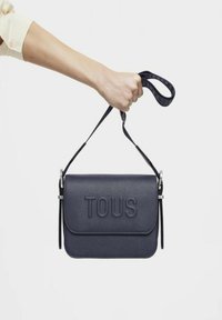 Bolso bandolera de piel navy con el logo "TOUS" en relieve, forma rectangular, correa ajustable y superficie texturizada suave.