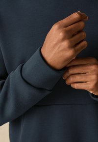 Tmavě modrý textilní materiál s detailním záběrem rukou, které svírají manžetu. Materiál vypadá hladce, s viditelnou texturou a jemnými detaily.
