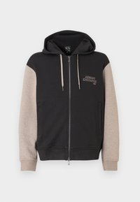Sudadera con capucha y cremallera, con cuerpo negro y mangas beige. El material es suave con una textura lisa. Logo de la marca en el pecho en un color contrastante.