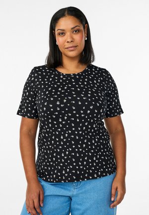 Mujer con cabello negro a la altura de los hombros, lleva una camiseta negra de manga corta con un patrón floral blanco y vaqueros azules, de pie contra un fondo blanco.