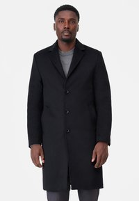 Svart ullrock med skräddarsydd design, enfaldig fram, fyra knappar och sidofickor. Bärs över en grå tröja.