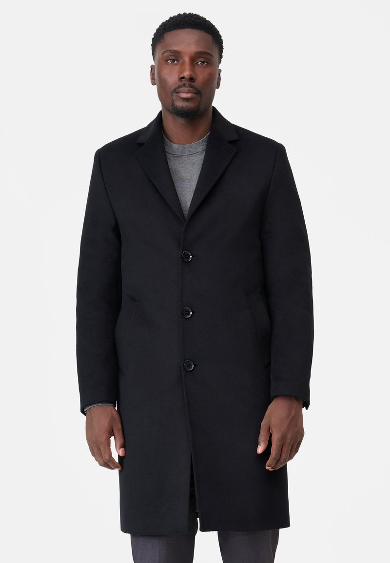 Svart ullrock med skräddarsydd design, enfaldig fram, fyra knappar och sidofickor. Bärs över en grå tröja.