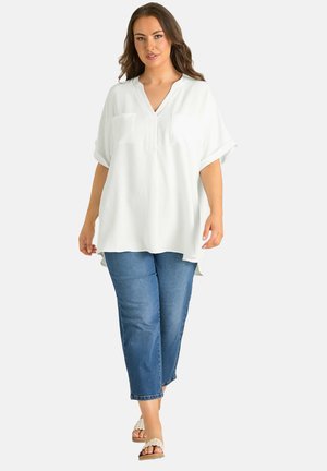 Kobieta idąca, ubrana w luźną białą bluzkę z krótkim rękawem, niebieskie spodnie typu cropped oraz beżowe sandały na wsuwkę na jasnym, jednolitym tle.