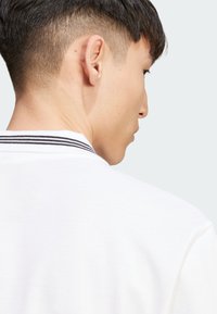 Witte poloshirt van een gestructureerde stof, met een zwart-wit gestreepte kraag. De focus ligt op de achterkant van de kraag.