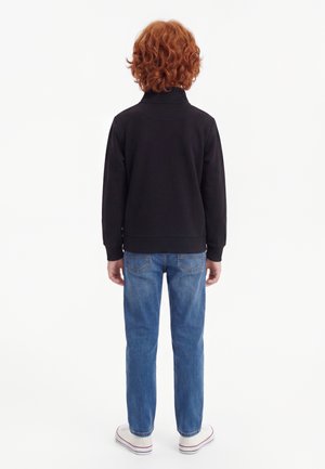 Enfant aux cheveux roux bouclés, debout de dos, portant une veste noire, un jean bleu et des baskets blanches sur fond blanc.