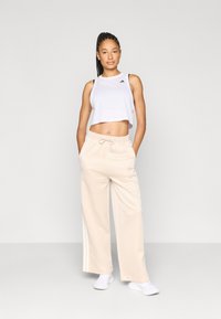 Bel top brez rokavov, ki je prirezan, in bež široke trenirke s širokim elastičnim pasom in vezalko. Hlače imajo bele stranske črte. Bele superge.