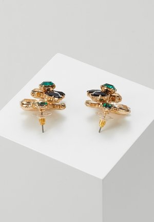 Boucles d'oreilles clous ton or avec des pierres facettées vertes et noires, montées en un design géométrique superposé sur un bloc d'exposition blanc.