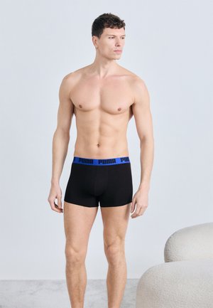 Νέος μυώδης άνδρας όρθιος, φοράει μαύρα εσώρουχα boxer της Puma με μπλε ελαστική ζώνη στη μέση, μπροστά σε απλό ανοιχτό γκρι φόντο.