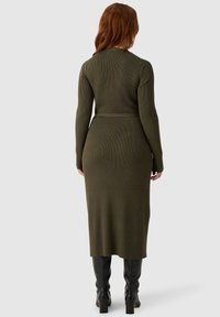 Olivgrünes, geripptes Strickkleid mit langen Ärmeln, tailliertem Schnitt und Midi-Länge, kombiniert mit schwarzen kniehohen Stiefeln. Glatte Textur.