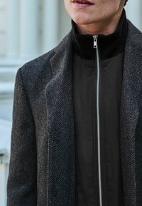 Gråtexturerad ytterjacka med ett mörkt innerlager. Har en hög krage och silverdragkedja, vilket visar på kontrasterande material och färger.