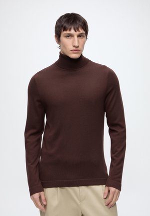 Junger Mann mit kurzen braunen Haaren, der einen eng anliegenden dunkelbraunen Rollkragenpullover und beige Hosen vor einem schlichten hellen Hintergrund trägt.