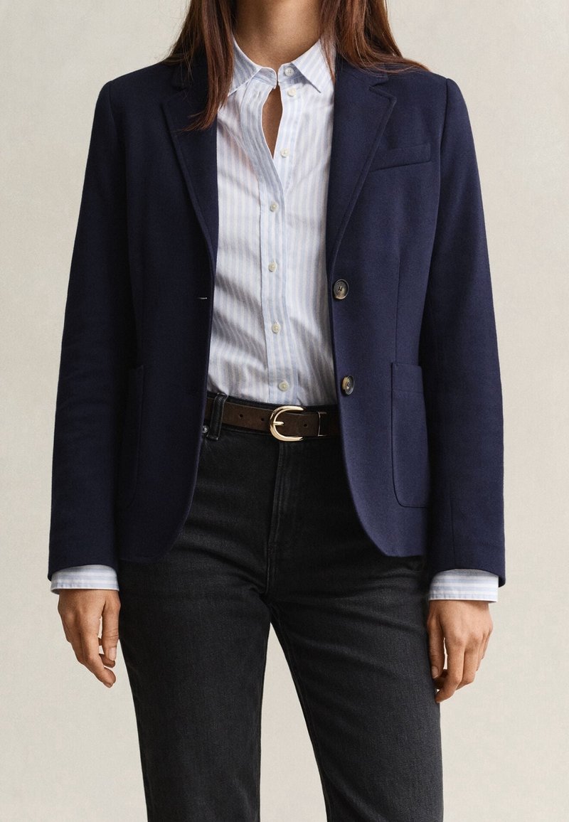 Femme portant un blazer bleu marine, une chemise à rayures bleu clair boutonnée, un pantalon noir et une ceinture marron avec une boucle dorée sur fond uni.