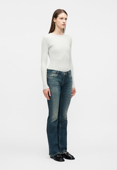 Γυναίκα που φοράει γκρι ανοιχτόχρωμο μακρυμάνικο εφαρμοστό τοπ, ξεβαμμένο μπλε τζιν με ίσια γρ αμμή και μαύρα μοκασίνια, στέκεται μπροστά σε λευκό φόντο.