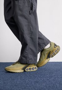 En person som bär olivgröna sneakers med synliga luftkuddar och mörkgrå byxor står på ett blått heltäckningsmatta.