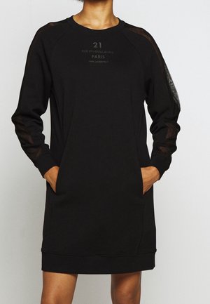 Femme portant une robe noire à manches longues avec des manches semi-transparentes et des poches avant, portant le texte « 21 Rue St-Guillaume PARIS » sur la poitrine.