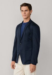 Muž v tmavomodrom saku cez modrú košeľu a bielych nohaviciach stojaci pred jednoduchým svetlosivým pozadím.