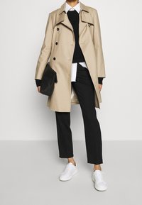 Beige trenčkot s dvojitými knoflíky přes černý svetr a bílou košili, spárovaný s černými kalhotami a bílými teniskami.