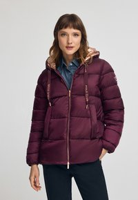 Burgundy pufferjacka med kviltat mönster, rosé-guld accenter och huva. Har dragkedjestängning och sidofickor. Slät yta.