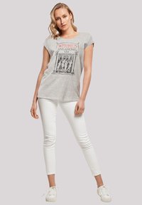 Grå t-shirt med korta ärmar som har en svart och röd grafik med texten "Häxor Lever Bland Oss!", tillsammans med vita skinny jeans och vita sneakers.