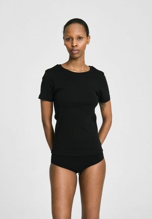 Giovane donna con maglietta nera a maniche corte e slip neri, in piedi con le mani dietro la schiena su uno sfondo neutro.