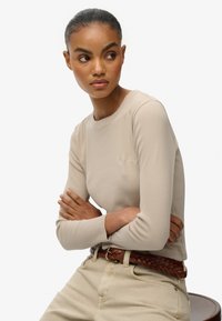 Beige langærmet top med rund halsudskæring, lavet af elastisk stof, med tekstureret broderi på brystet og en figursyet silhuet.