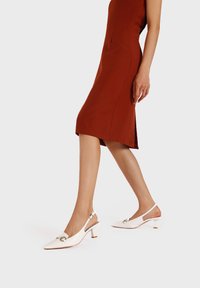 Scarpe slingback bianche in vernice con punta affusolata, piccolo tacco a blocco e dettagli dorati. Texture liscia e design minimalista.