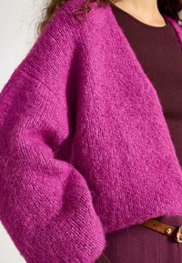 Oversized cardigan i levende fuchsia, strikket tekstur, med løse ærmer og en blød finish. Båret over en brun top med bælte.