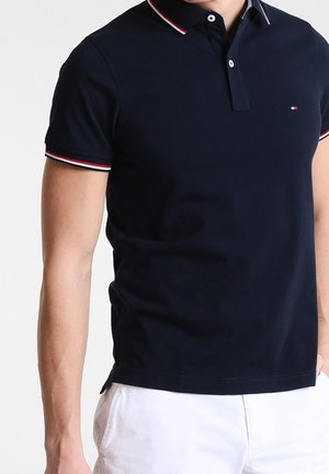 Mand iført en navyblå polo med rød, hvid og navy trim på krave og ærmer, kombineret med hvide shorts.